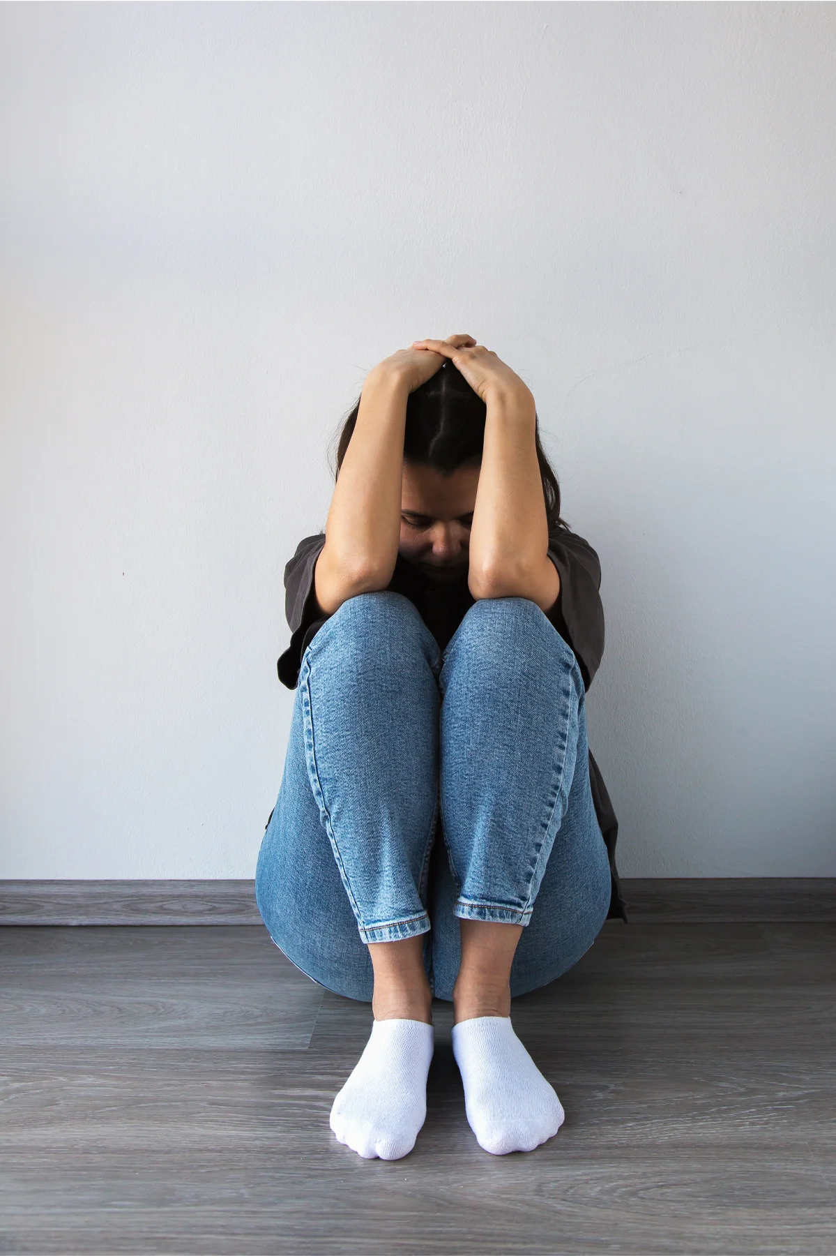 A woman sitting on the floor against a wall, hugging her knees and holding her head in distress, symbolizing depression, anxiety, and emotional struggle, vertical photo