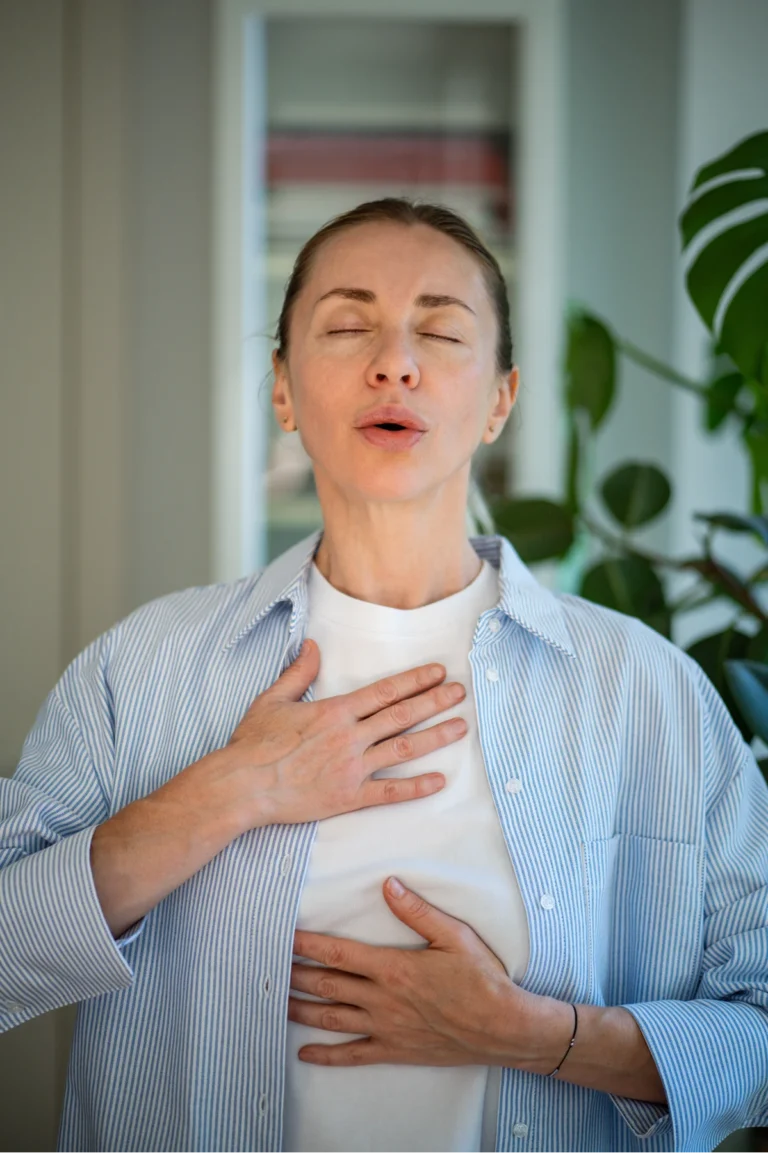 Calm middle aged woman practicing breathing meditation techniques standing with closed eyes, self caring at home. Focused on mental health female holding hands on chest and belly doing pranayama yoga.