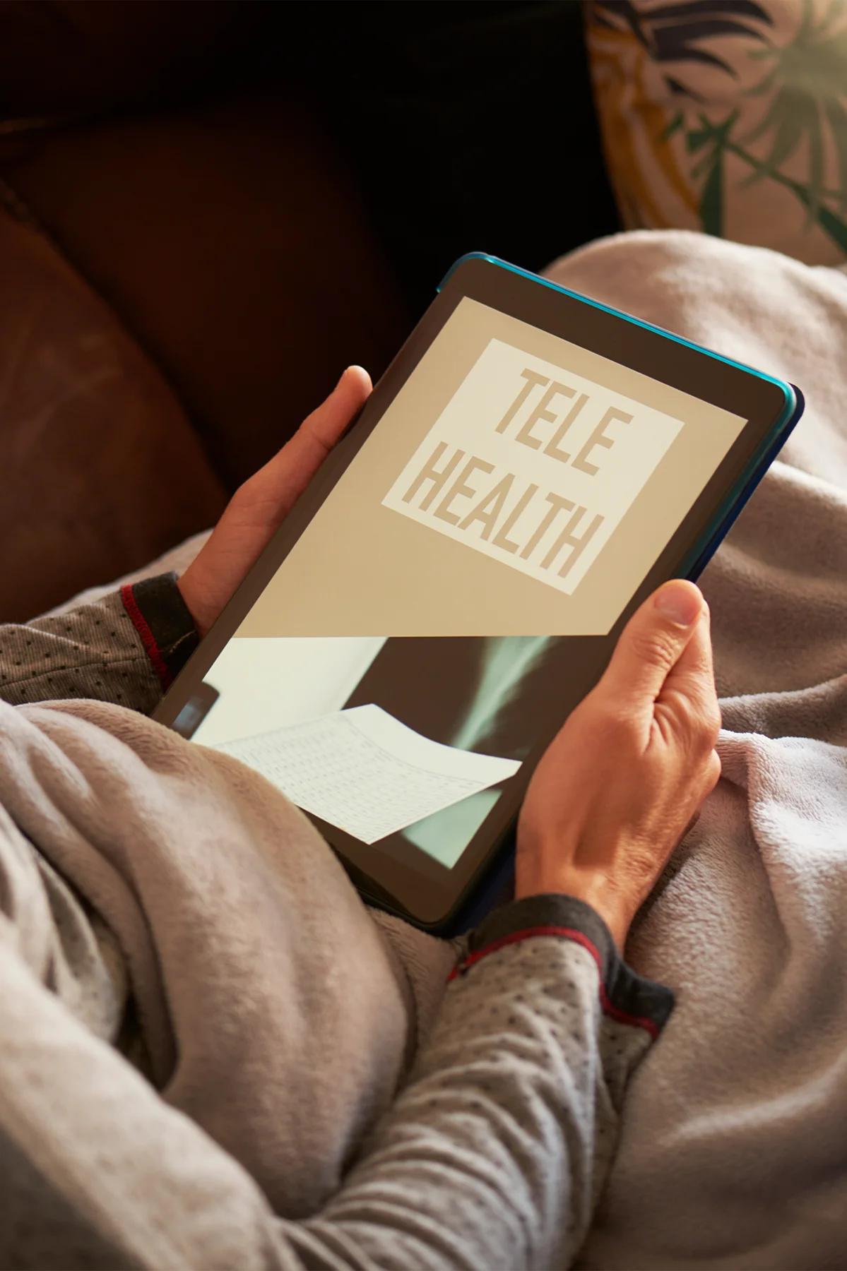 a young caucasian man, lying comfortably in a sofa wrapped in a warming gray blanket, has his tablet in his hands with the text telehealth on its screen
